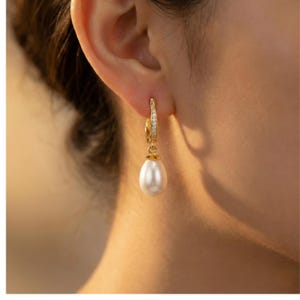 May include: A gold hoop earring with small clear stones and a dangling white teardrop-shaped pearl. The earring is shown on a person's ear, highlighting the jewelry's design and elegance.