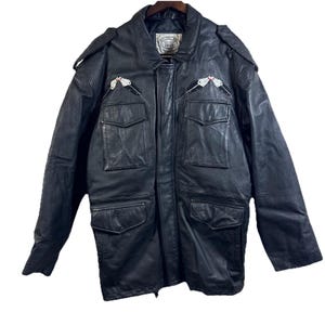 Peut inclure: Veste en cuir noir avec plusieurs poches et des écussons de pistolets décoratifs. La veste a un col et des épaulettes sur les épaules. La veste est suspendue à un cintre en bois.