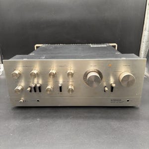 Pode incluir: Um amplificador estéreo Pioneer modelo SA-9800 prateado com painel frontal de metal escovado. A unidade possui vários botões circulares para graves, agudos, volume e função, juntamente com vários interruptores e uma entrada para auscultadores.