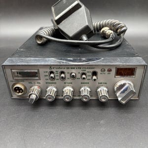 May include: A black Cobra 29 NW LTD Classic CB radio with a silver front panel, featuring multiple control knobs, toggle switches, a digital channel display, a signal meter, and a black handheld microphone with a coiled cord resting on top.
