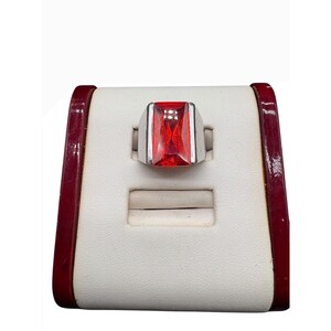 May include: A silver ring with a large, rectangular, red gemstone. The ring is displayed on a white and burgundy jewelry display stand. The gemstone is the focal point, catching the light.
