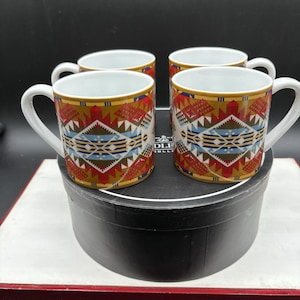 May include: A set of four ceramic mugs featuring a repeating geometric pattern in shades of mustard yellow, red, blue, and white. Each mug has a white interior and a white handle, arranged in a square formation on a black circular container.
