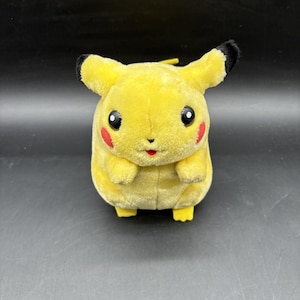 Puede incluir: Un peluche amarillo con cuerpo redondeado, dos orejas puntiagudas con puntas negras, dos brazos pequeños y dos pies. La cara tiene dos ojos negros con reflejos blancos, una nariz pequeña, una boca diminuta y dos parches circulares rojos en las mejillas.
