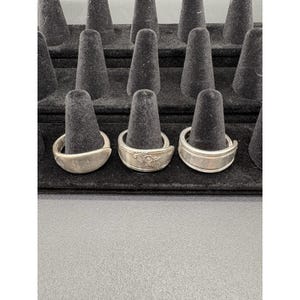 May include: Silver spoon rings displayed on a black velvet ring holder. Three rings are visible, each with a unique design. One ring has a smooth, curved band, while another features a floral pattern. The third ring has a simple, ridged design.