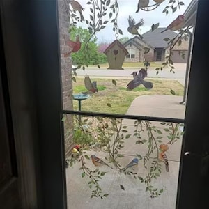 May include: A glass door decorated with bird and leaf decals. The decals include various colorful birds, green leaves, and a small birdhouse. The scene outside the door includes a bird bath, a house, and a tree.