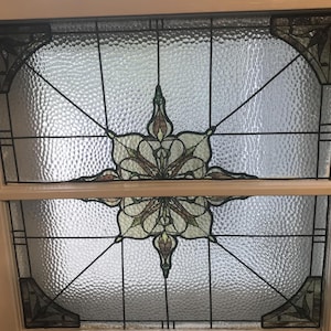May include: Stained glass window with a white frame. The window features a central floral design in shades of brown, green, and cream, with radiating lines and geometric patterns. The glass has a textured, bubbly appearance.