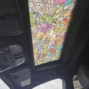 May include: A car sunroof featuring a colorful stained-glass design. The artwork depicts flowers, leaves, and a bird in shades of pink, yellow, green, and blue. The sunroof is framed by a black interior.