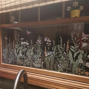 May include: A window with a floral design featuring various wildflowers and butterflies in shades of green, pink, and blue. The window is framed in wood and has a bamboo shade above. A small yellow pot with white flowers sits on the shelf above the window.