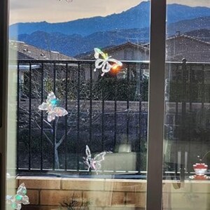 May include: A window scene featuring several decorative butterfly ornaments. The butterflies are iridescent, with a shimmering effect, and are attached to the window. The background includes a black metal fence, brickwork, and a view of distant mountains.