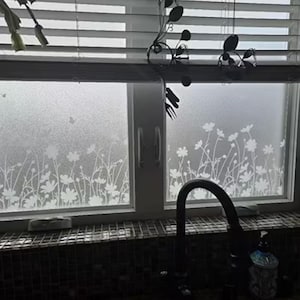 May include: Frosted window film with a white floral design. The film is applied to a double-pane window, with a dark faucet and blinds in the background. The floral pattern includes daisies and other wildflowers.