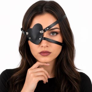 Könnte beinhalten: Eine schwarze herzförmige Augenklappe aus glattem Material mit dünnen, verstellbaren Riemen, die um den Kopf verlaufen. Die Klappe ist mit silberfarbenen Metallnieten und Beschlägen befestigt und bedeckt ein Auge mit einer Herzsilhouette.