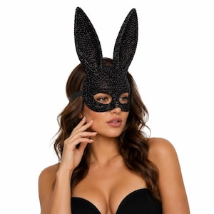 Op de afbeelding: Een zwart konijnenmasker met lange, rechtopstaande oren en een getextureerd, glinsterend oppervlak dat het bovenste deel van het gezicht bedekt. Het masker heeft uitsparingen voor de ogen en wordt vastgezet met een dunne zwarte band.