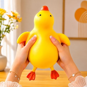 May include: A bright yellow squeeze toy shaped like a duck with a smooth, rounded body, small black eyes, and a distinct orange beak and webbed feet. The toy has a soft, pliable texture and is held firmly on both sides, showcasing its flexible form.