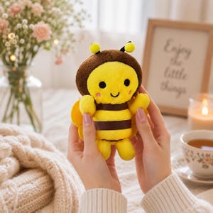 May include: A plush bee toy with a yellow and brown striped body, a brown head, small black eyes, a smiling mouth, and pink cheeks. The soft toy features yellow wings on the sides and small yellow arms and legs, crafted from a fuzzy fabric.