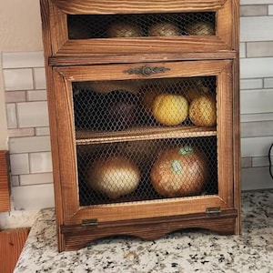 May include: A rustic wooden storage cabinet with a mesh front, filled with various vegetables. The cabinet has a warm brown finish and is divided into two sections. The cabinet is sitting on a speckled countertop.