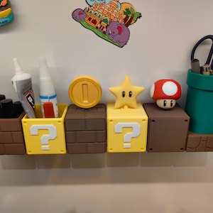 May include: A collection of novelty desk organizers and containers in the style of the Super Mario Bros. video game. Includes a brown brick block, yellow question mark blocks, a coin, a star, a mushroom, and a green pipe. Some containers hold pens, scissors, and glue.