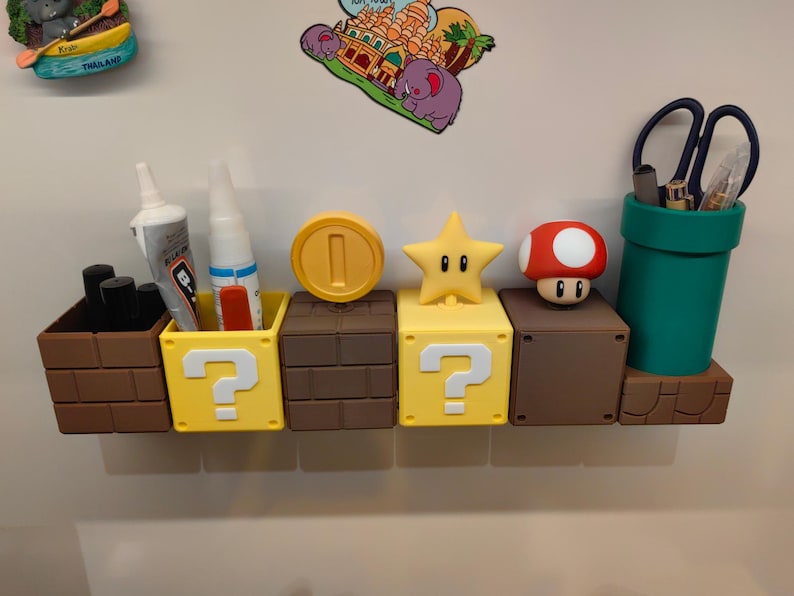 May include: A collection of novelty desk organizers and containers in the style of the Super Mario Bros. video game. Includes a brown brick block, yellow question mark blocks, a coin, a star, a mushroom, and a green pipe. Some containers hold pens, scissors, and glue.