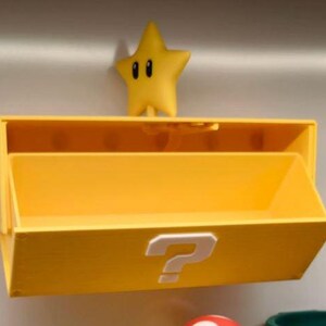 May include: A yellow storage box with a question mark on the front, and a yellow star figure on top. The box has an open compartment and is designed to be mounted on a wall. The star has black eyes.