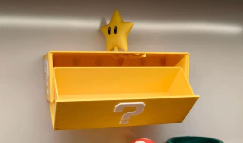 May include: A yellow storage box with a question mark on the front, and a yellow star figure on top. The box has an open compartment and is designed to be mounted on a wall. The star has black eyes.