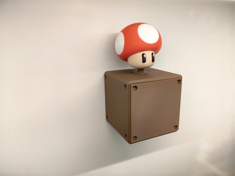 May include: A brown square block with a red mushroom character on top. The mushroom has a white-spotted red cap and a neutral face. The block has four small circular indentations on each side.