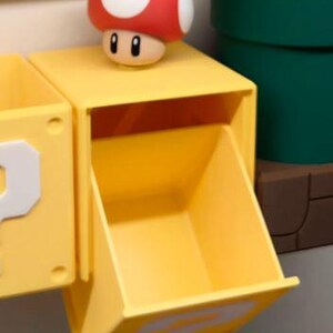 May include: A yellow storage box with a question mark on the side, a red and white mushroom figure, and a green pipe. The box has an open drawer. The mushroom figure is on top of the box.