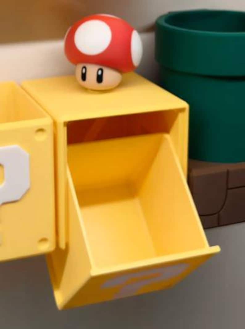 May include: A yellow storage box with a question mark on the side, a red and white mushroom figure, and a green pipe. The box has an open drawer. The mushroom figure is on top of the box.