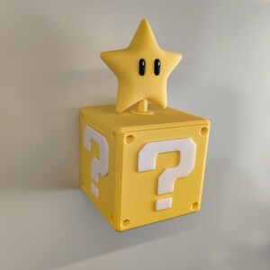 May include: A yellow, square block with white question marks on two sides, topped with a yellow star with black eyes. The block and star are likely inspired by a video game.