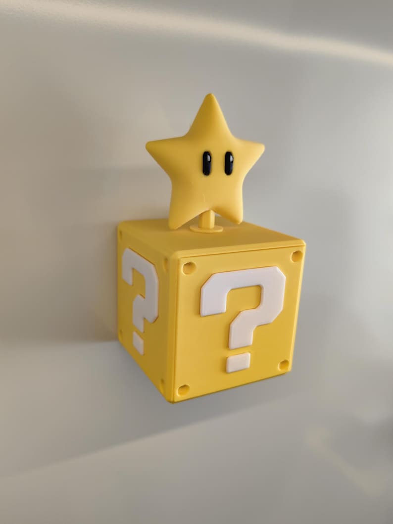 May include: A yellow, square block with white question marks on two sides, topped with a yellow star with black eyes. The block and star are likely inspired by a video game.