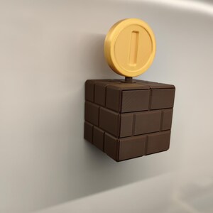 May include: A brown brick block with a yellow coin on top, reminiscent of the Super Mario video game series. The coin has a number 1 in the center. The block is made of several rectangular bricks.