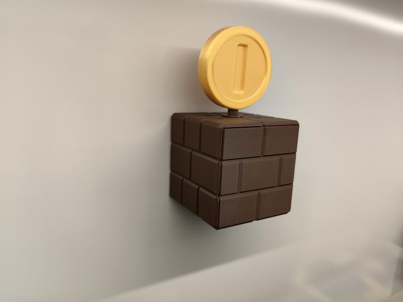 May include: A brown brick block with a yellow coin on top, reminiscent of the Super Mario video game series. The coin has a number 1 in the center. The block is made of several rectangular bricks.