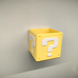 May include: A yellow, square box with a white question mark on each side. The box is a solid color and appears to be made of plastic. The box is a decorative item.