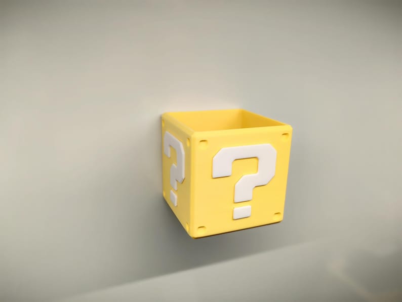 May include: A yellow, square box with a white question mark on each side. The box is a solid color and appears to be made of plastic. The box is a decorative item.