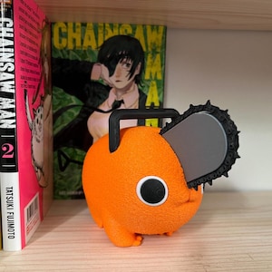 May include: An orange Pochita figure from the anime series Chainsaw Man, with a black handle and chainsaw blade. The figure is on a shelf with manga volumes and a Chainsaw Man poster in the background.