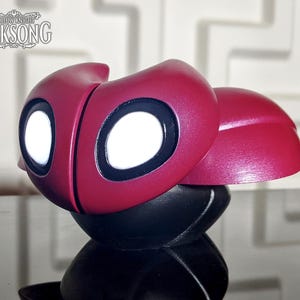 May include: A red and black figurine with large white eyes is shown in close-up. The figurine has a glossy finish and is set against a blurred background. The text "SILKSONG" is visible in the upper left corner.