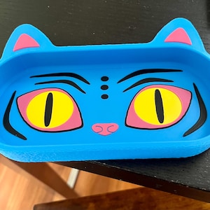 Puede incluir: Una bandeja azul con forma de gato con orejas y nariz rosas, y ojos amarillos con pupilas negras. La bandeja tiene un diseño de dibujos animados, perfecta para guardar objetos pequeños. La bandeja está hecha de un material flexible.