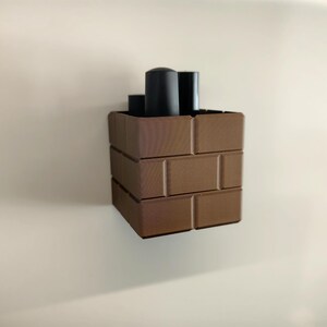 May include: A brown, brick-patterned container with three black cylindrical objects inside. The container is made of rectangular blocks, creating a stacked appearance. The background is a plain, off-white wall.