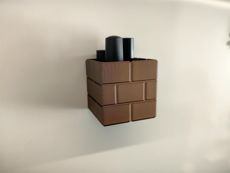 May include: A brown, brick-patterned container with three black cylindrical objects inside. The container is made of rectangular blocks, creating a stacked appearance. The background is a plain, off-white wall.