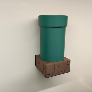 May include: A teal green pipe, reminiscent of a video game, is mounted on a brown, stone-like base. The cylindrical pipe has a wide rim and sits atop a square base with a textured design. The items are mounted on a white wall.