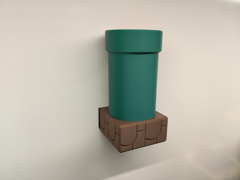 May include: A teal green pipe, reminiscent of a video game, is mounted on a brown, stone-like base. The cylindrical pipe has a wide rim and sits atop a square base with a textured design. The items are mounted on a white wall.