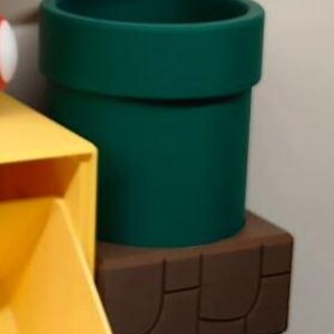 May include: A dark green, cylindrical pipe, reminiscent of a video game, sits atop a brown, rectangular base. The pipe has a wide opening and a thick rim. The base has a textured, brick-like design.