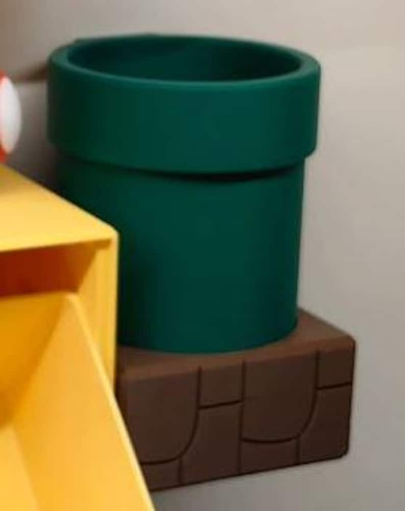 May include: A dark green, cylindrical pipe, reminiscent of a video game, sits atop a brown, rectangular base. The pipe has a wide opening and a thick rim. The base has a textured, brick-like design.