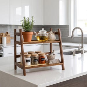 Op de afbeelding: Een houten plank met twee niveaus en geweven inzetstukken, met een potplant, glazen theepot en potten met de labels "Zout", "Peper" en "Paprika". Een vijzel en stamper en een kleine theepot staan ook op de plank. De plank staat op een wit aanrecht.