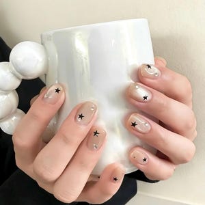 Puede incluir: Uñas manicuradas con base nude, adornadas con calcomanías de estrellas negras y pequeños diamantes de imitación. Las uñas son cortas y cuadradas. Las manos sostienen una taza blanca con un asa esférica.