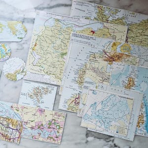 Op de afbeelding: Een verzameling papieren kaarten en kaart-thema uitsparingen met diverse regio's van Europa en de wereld. Het assortiment bevat rechthoekige vellen, ronde labels en geschulpte vormen, allemaal met gedetailleerde cartografische illustraties.