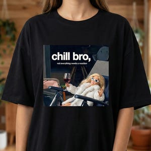 Op de afbeelding: Zwart T-shirt met een afbeelding van een pop met een zonnebril en een witte bontjas, die een glas wijn vasthoudt. De tekst "chill bro, not everything needs a reaction" staat boven de afbeelding.