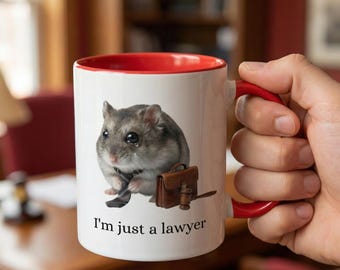 May include: White ceramic mug with a red interior and handle. The mug features a crying hamster wearing a tie and holding a briefcase. The text "I'm just a lawyer" is printed below the hamster.