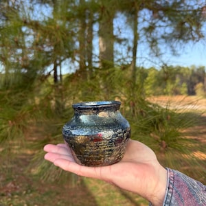 May include: A small, handcrafted ceramic vase with a textured surface. The vase has a dark, metallic glaze with shades of black, bronze, and green. The vase is held in a person's hand against a blurred outdoor background.