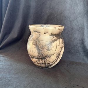 May include: A handcrafted, round vase with a light tan base and intricate black lightning patterns. The vase has a wide, slightly flared opening and a rounded body. The unique design adds a touch of rustic elegance to any home decor.