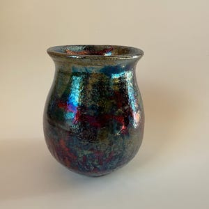 May include: A handmade ceramic vase with a unique iridescent glaze. The vase features a rounded body and a slightly flared rim. The glaze has a mix of colors, including blue, red, and green, creating a shimmering effect. The vase is a decorative item.