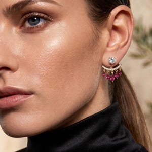Pode incluir: Um brinco ear jacket em tom dourado com um pino redondo transparente na frente e uma barra curva atrás do lóbulo. A barra é adornada com pequenas pedras transparentes e cinco pedras fúcsia em lapidação marquise pendentes em franja.